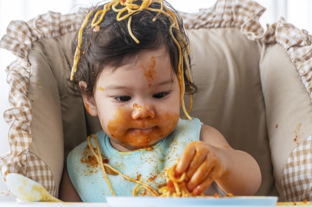 Adorable little child funny girl eating spaghetti with spoon while sitting in high-powered chair at home. Toddler child with tomato sauce making mess her face looking at parent. Self-feeding concept