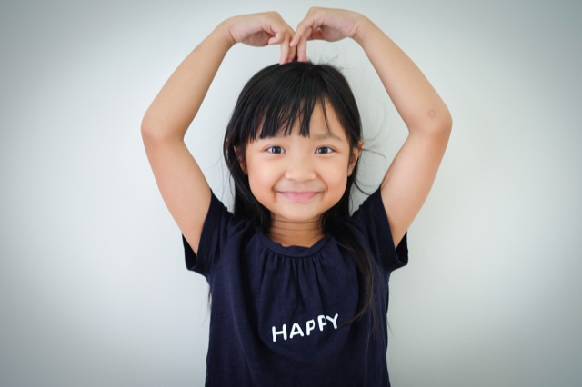 Portrait Asian child girl of smiling resting wearing dark blue t- shirt and pose making heart shape symbol with her arms